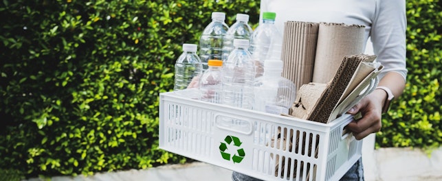 Kobieta trzymająca w dłoniach koszyk ze znakiem recyklingu, wypełniony pustymi opakowaniami tekturowymi i plastikowymi.